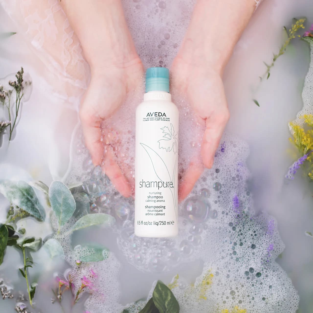 Aveda Shampure shampoo held above a bath of bubbles and botanical sprigs.